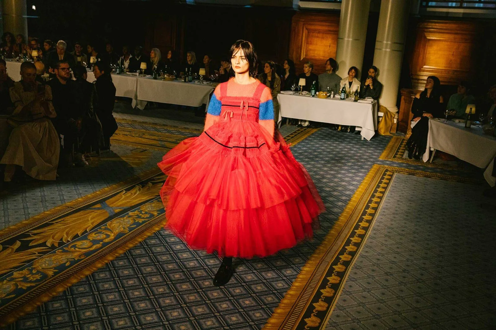 A model in a red tiered gown on a patterned carpet, a seated audience behind.