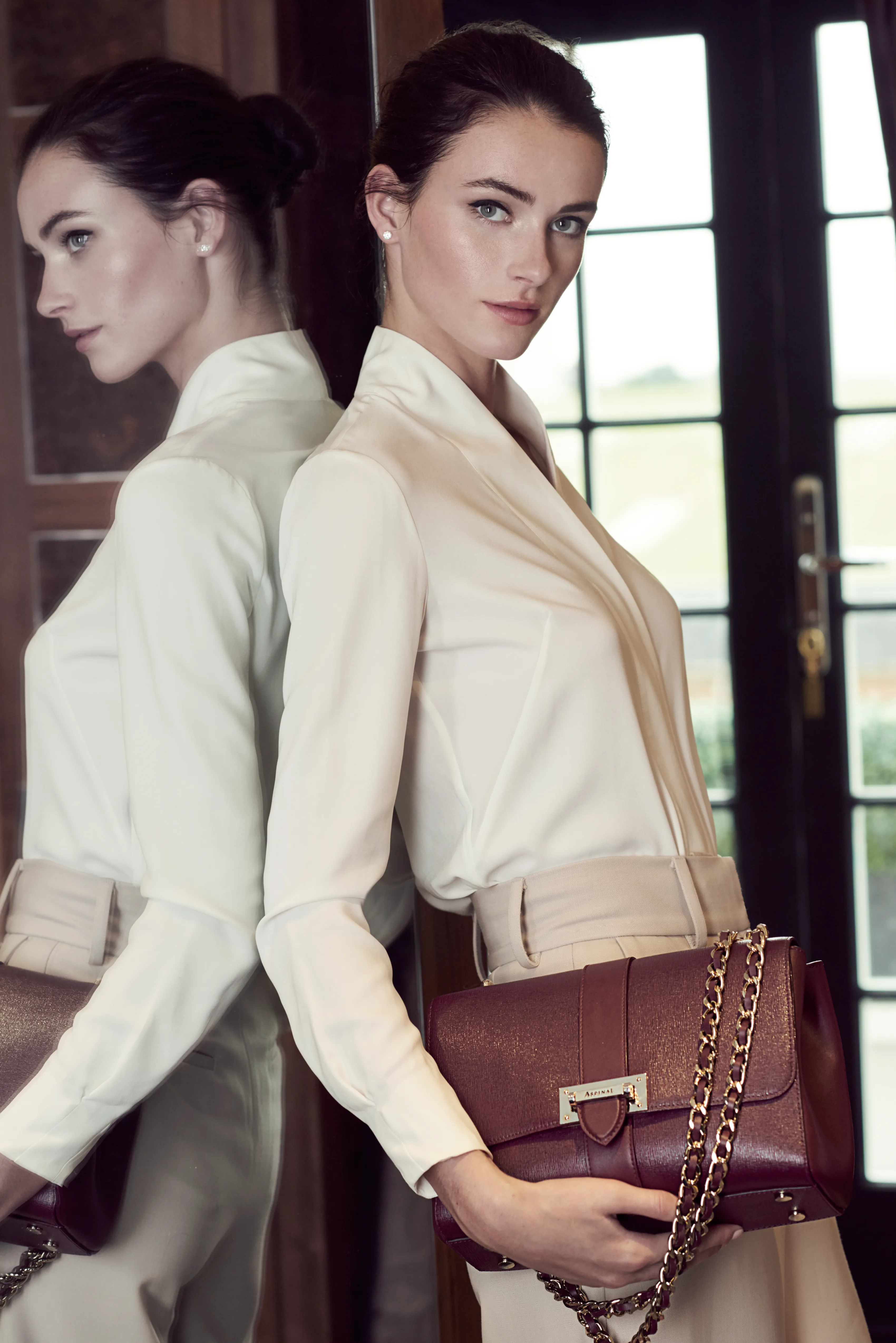 Two women in cream shirts at a tall window, one carrying a burgundy chain bag.