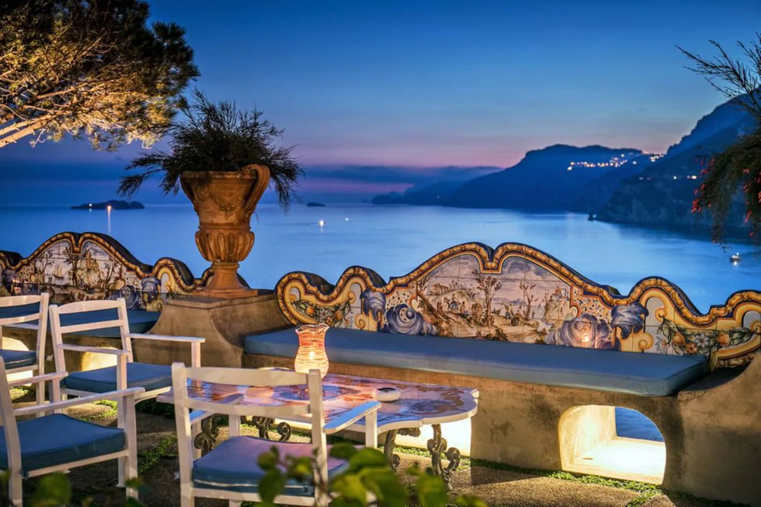 A clifftop terrace laid for dinner at dusk, above a bay.