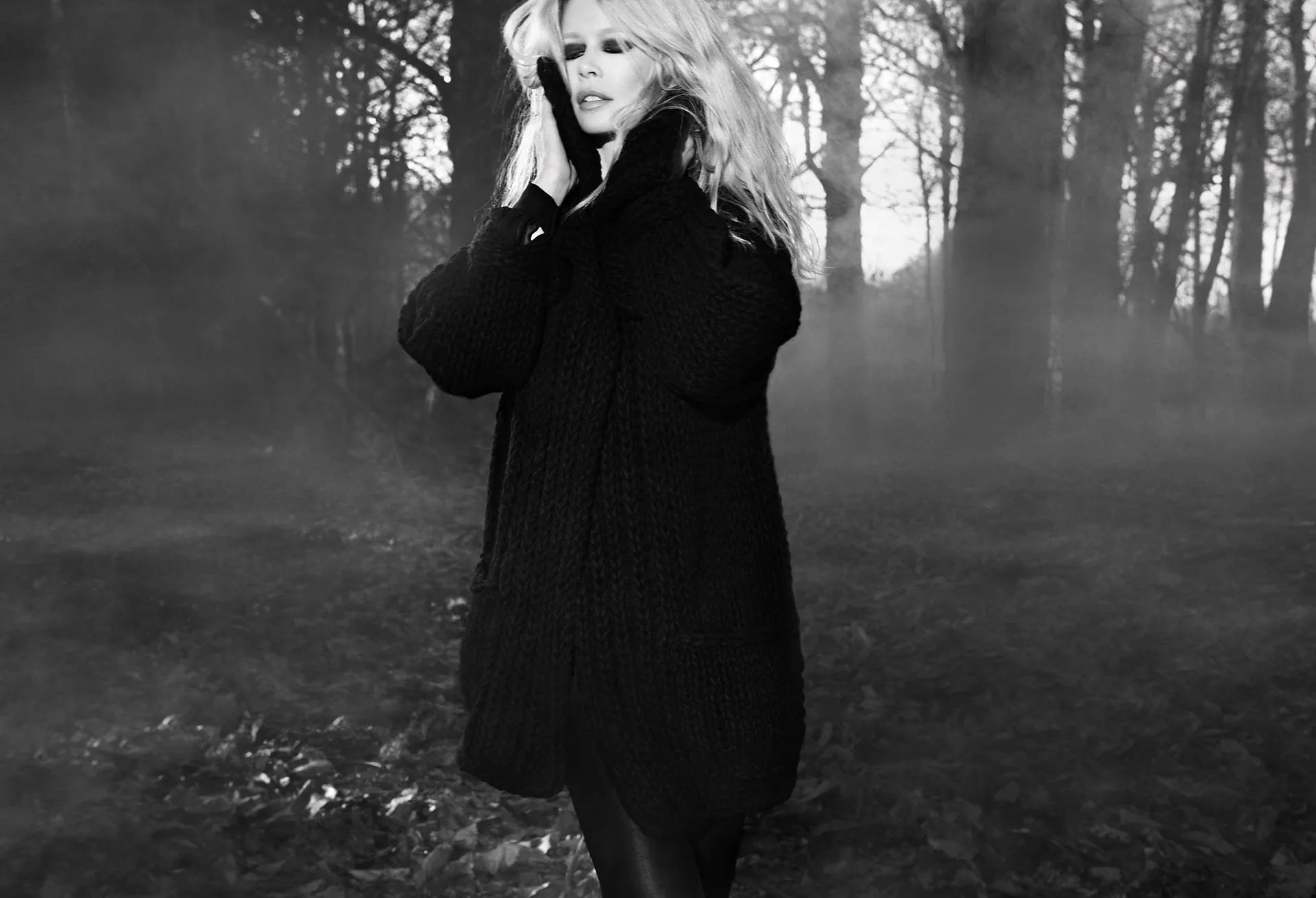A figure in a long dark coat in misty woodland, in black and white.