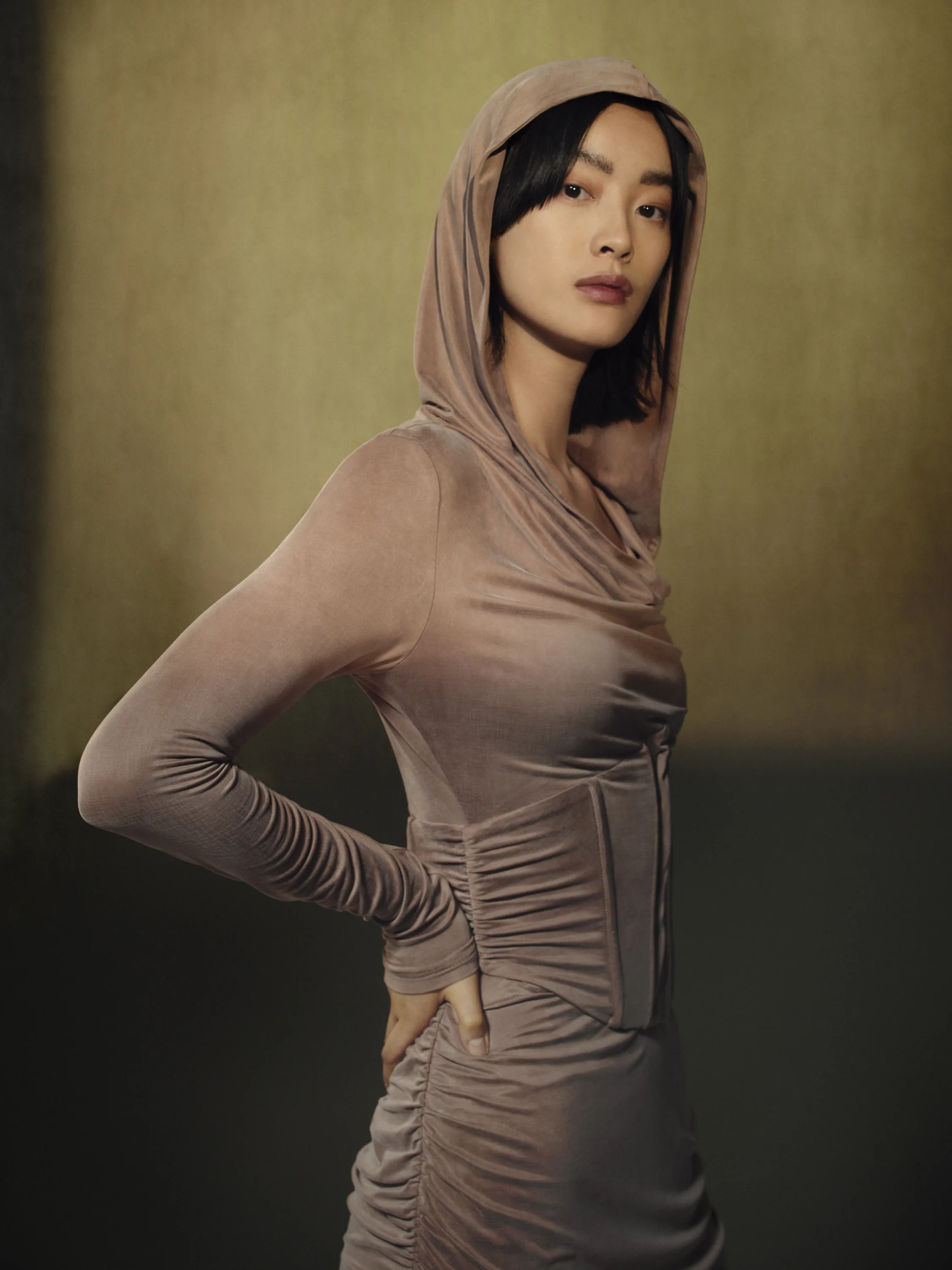 A model in a hooded mesh dress, against an olive backdrop.