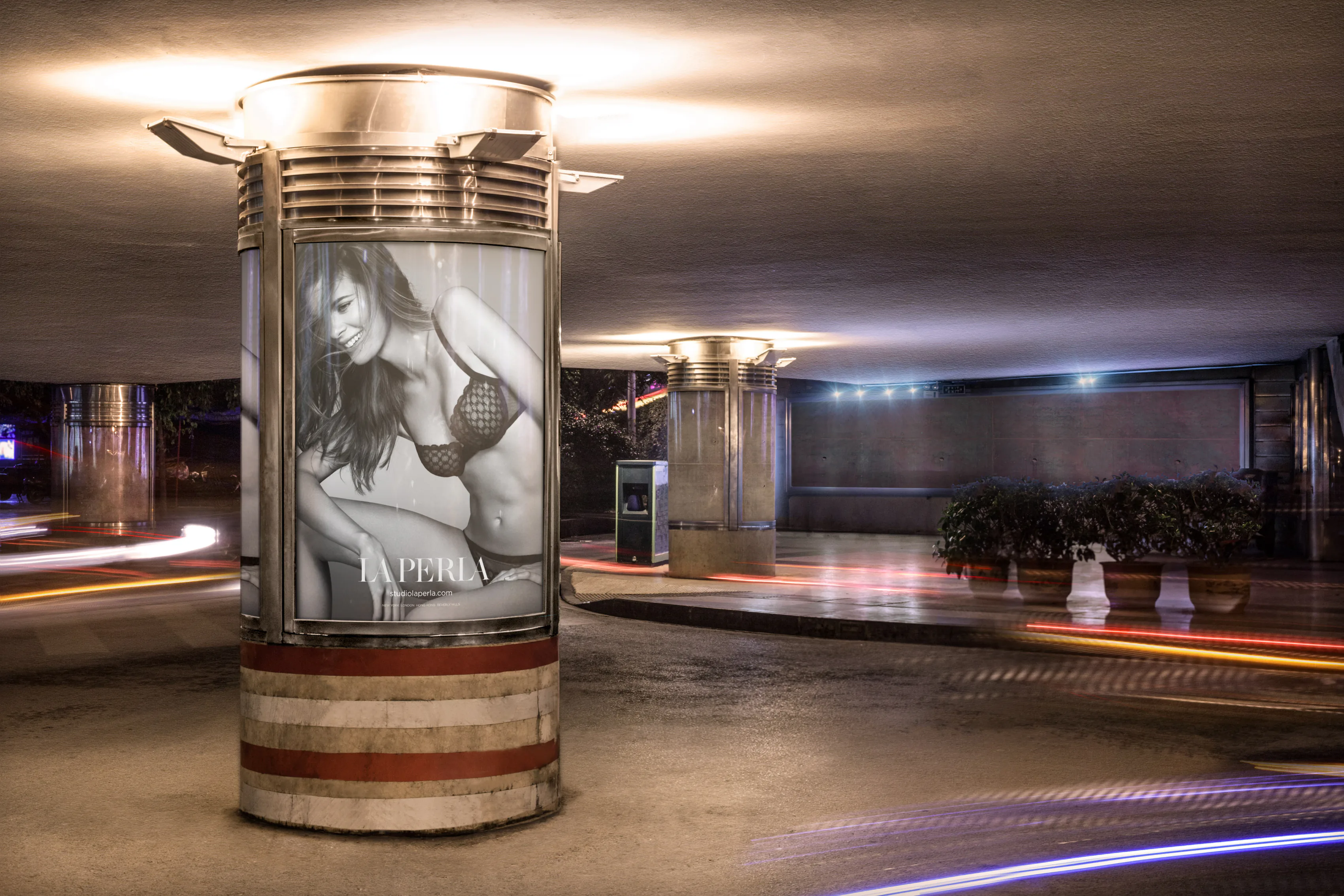 A lit advertising column carrying a La Perla poster under a road bridge, with traffic light trails running past it.