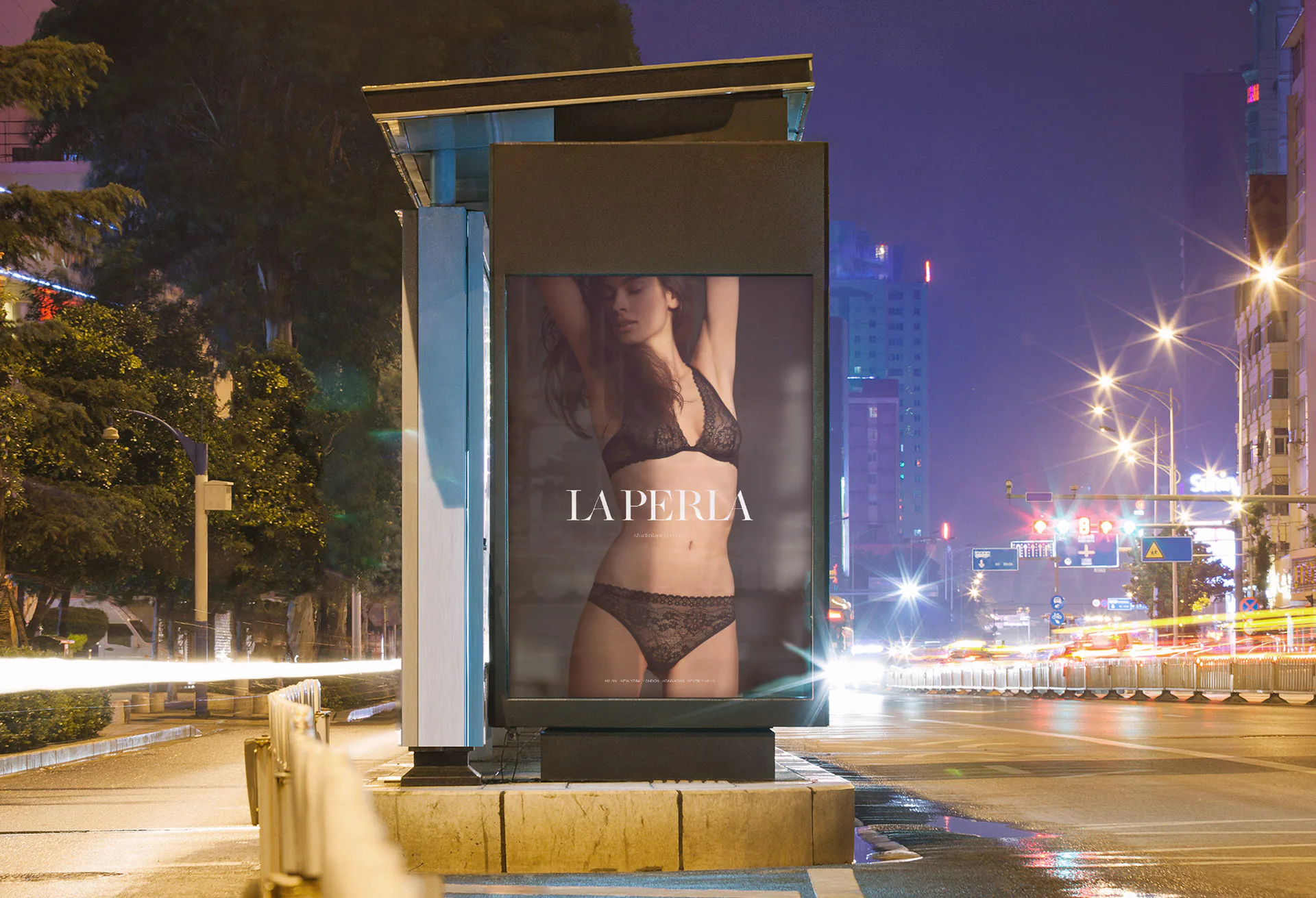 A La Perla poster lit in a bus shelter on a city street at night, with traffic light trails alongside it.