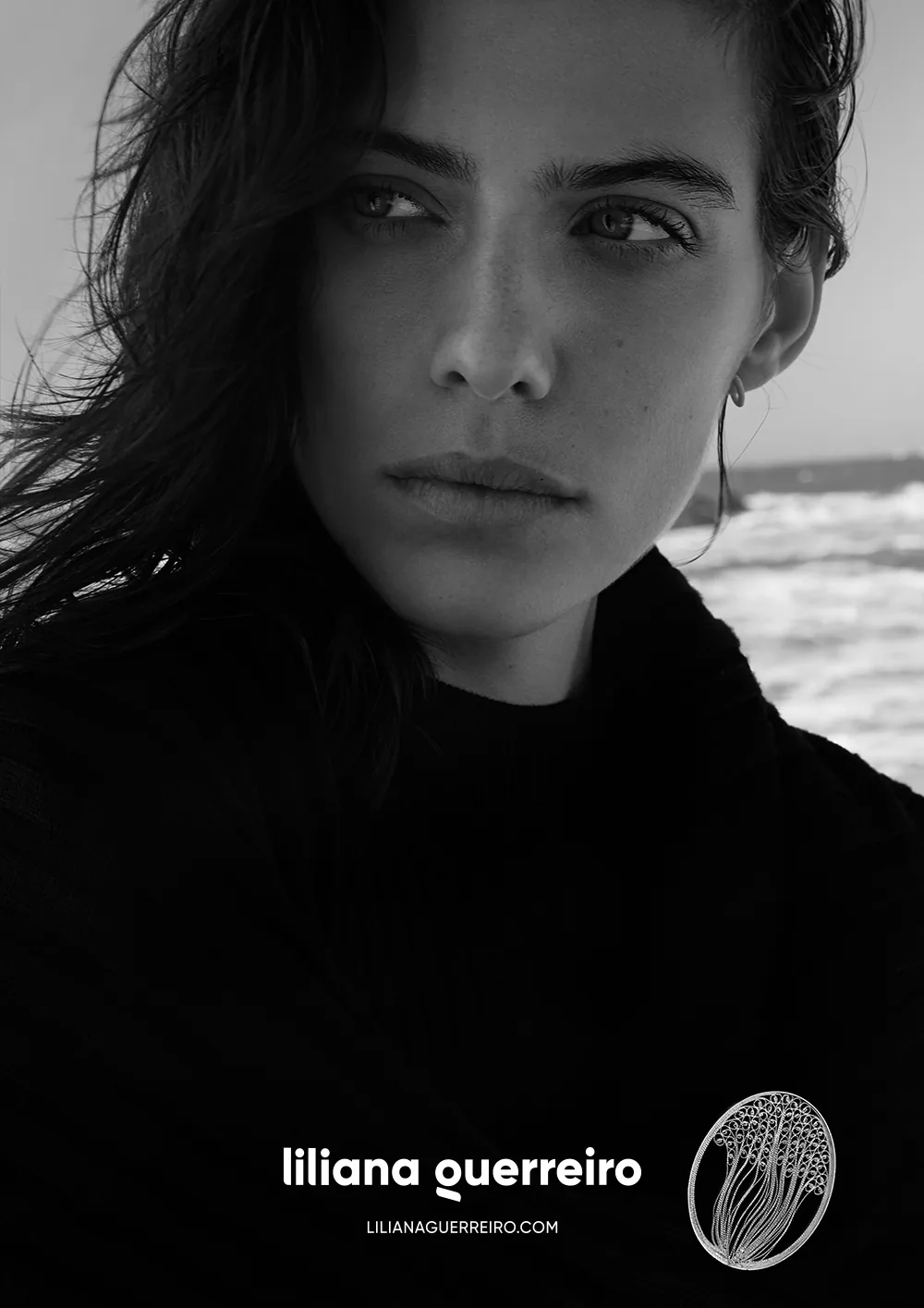 A black and white campaign portrait by the sea, the logotype and a filigree pendant below it.