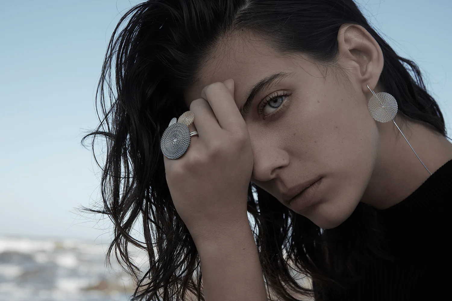A close portrait, one hand raised to the forehead, wearing a disc ring and matching earrings.