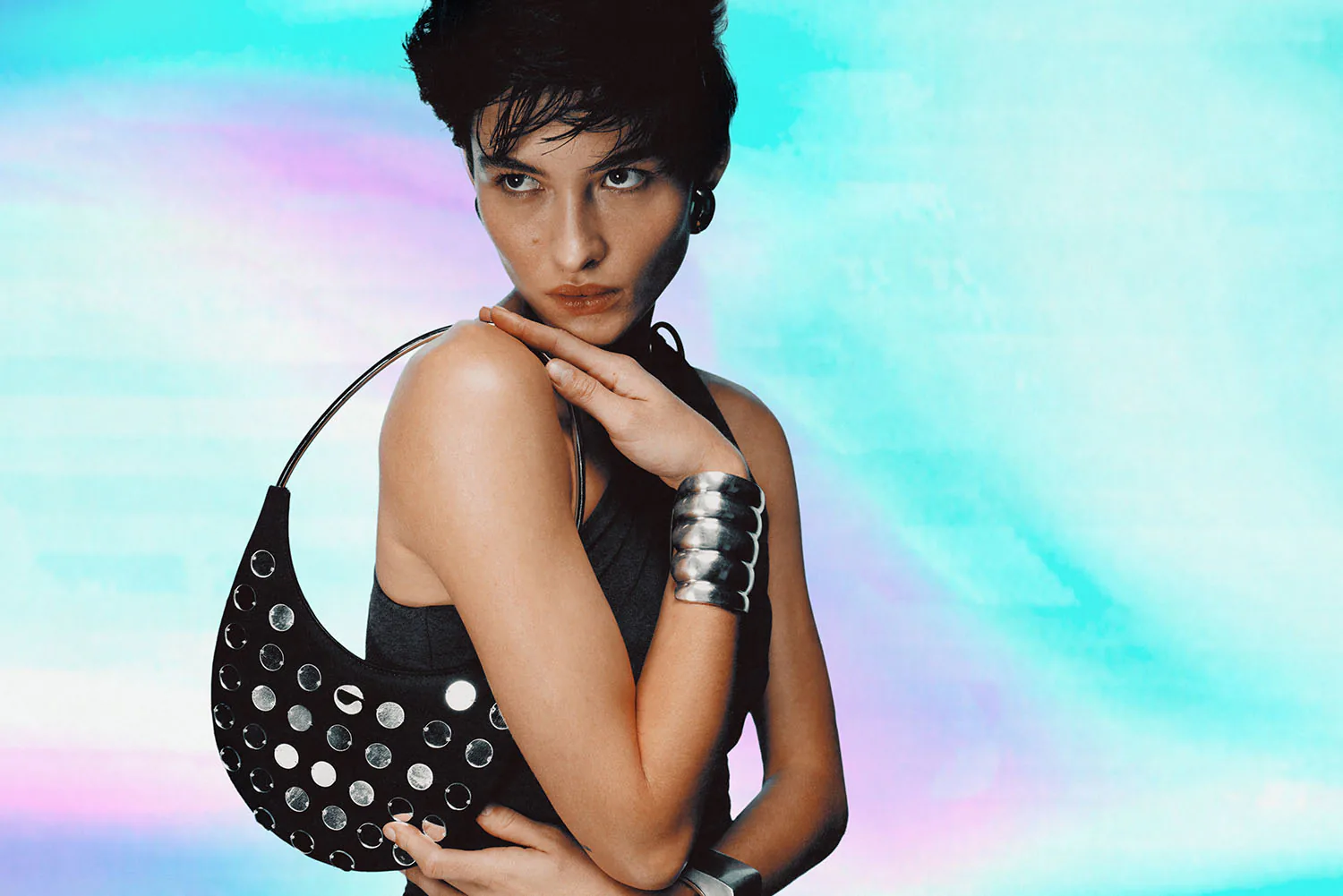 A model in a studded black dress and cuffs against an iridescent backdrop.