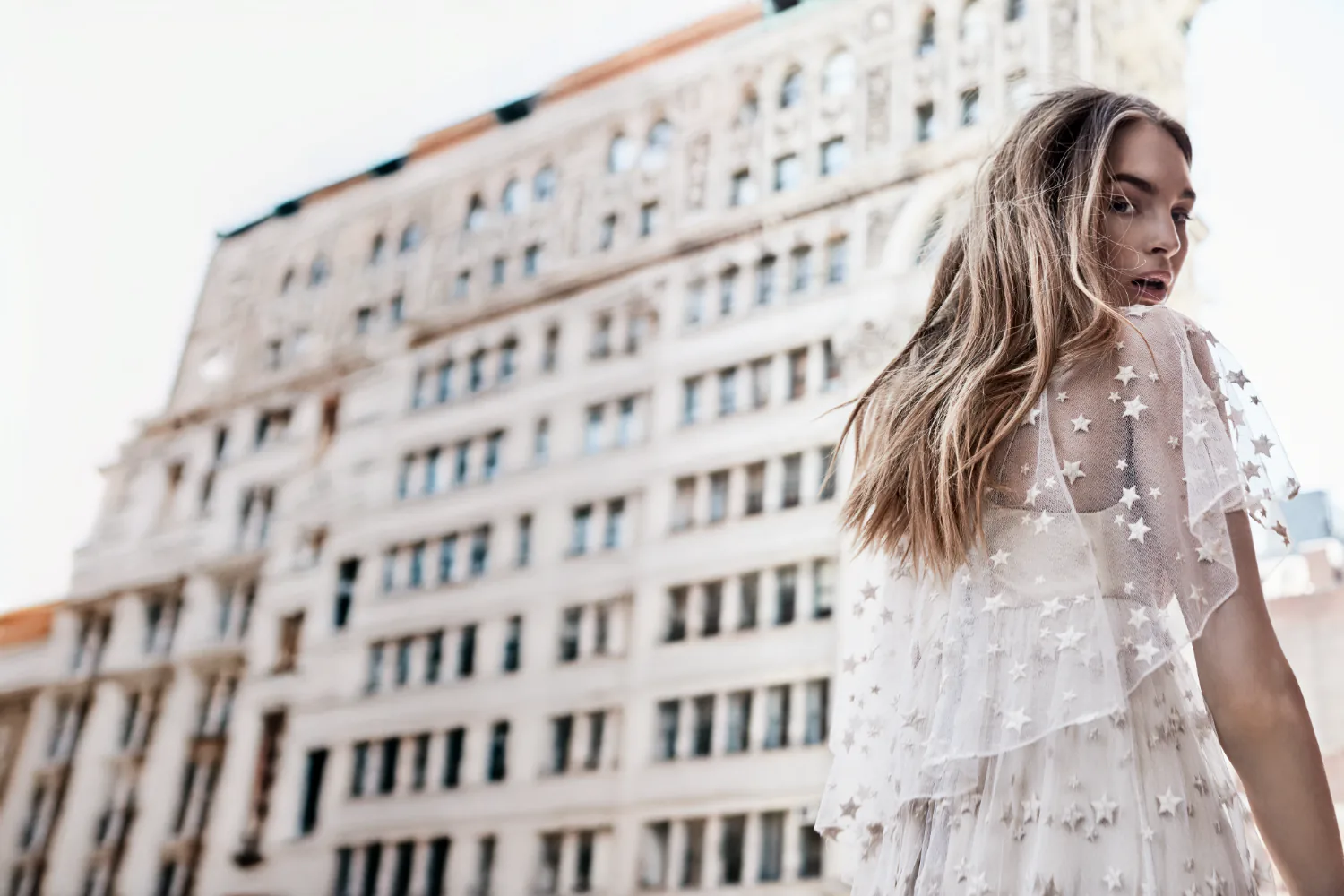 A model in a star-flocked sheer dress on a rooftop, a pale ornate building behind her.
