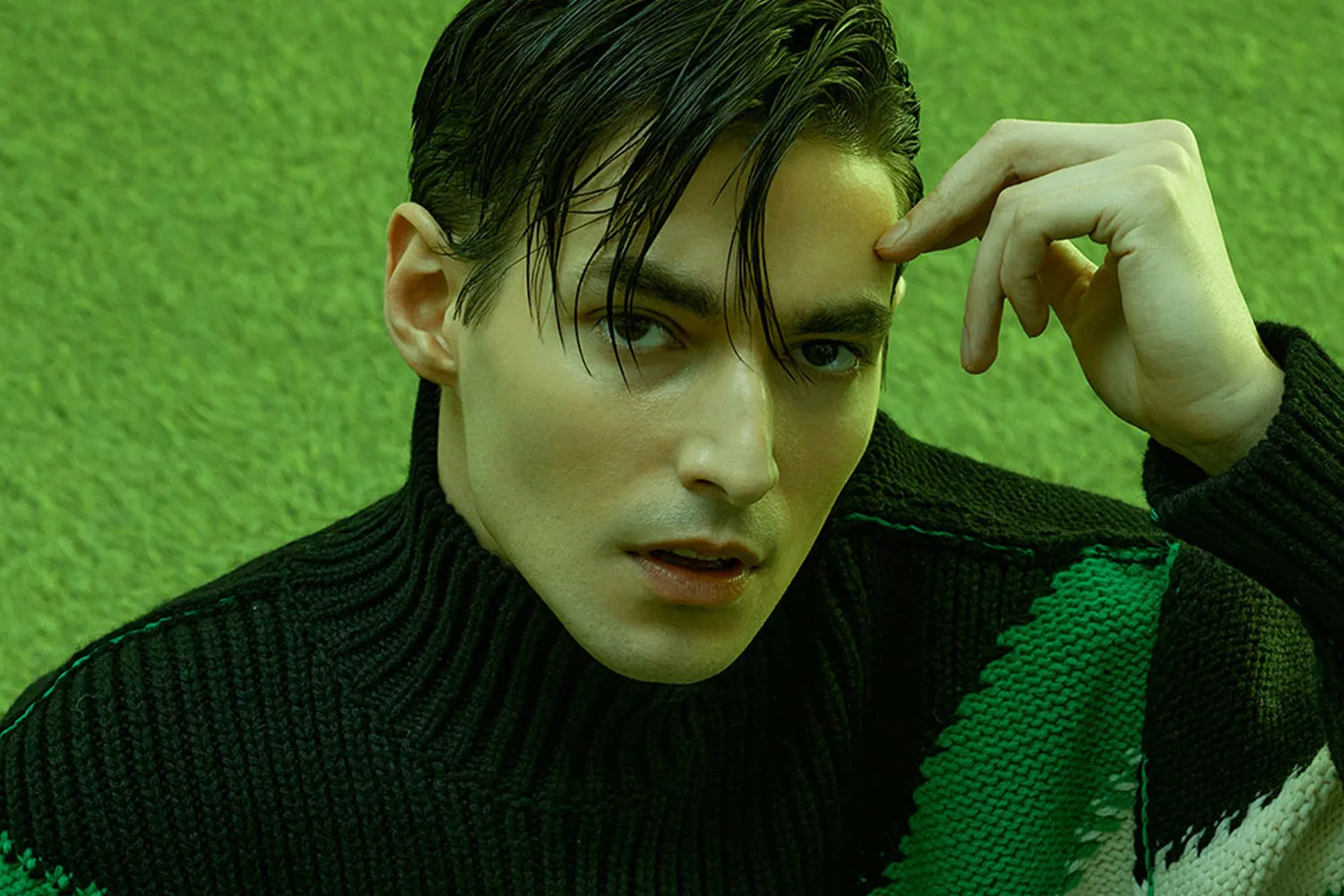 A man in a green knitted polo neck under green light, one hand raised to his temple.